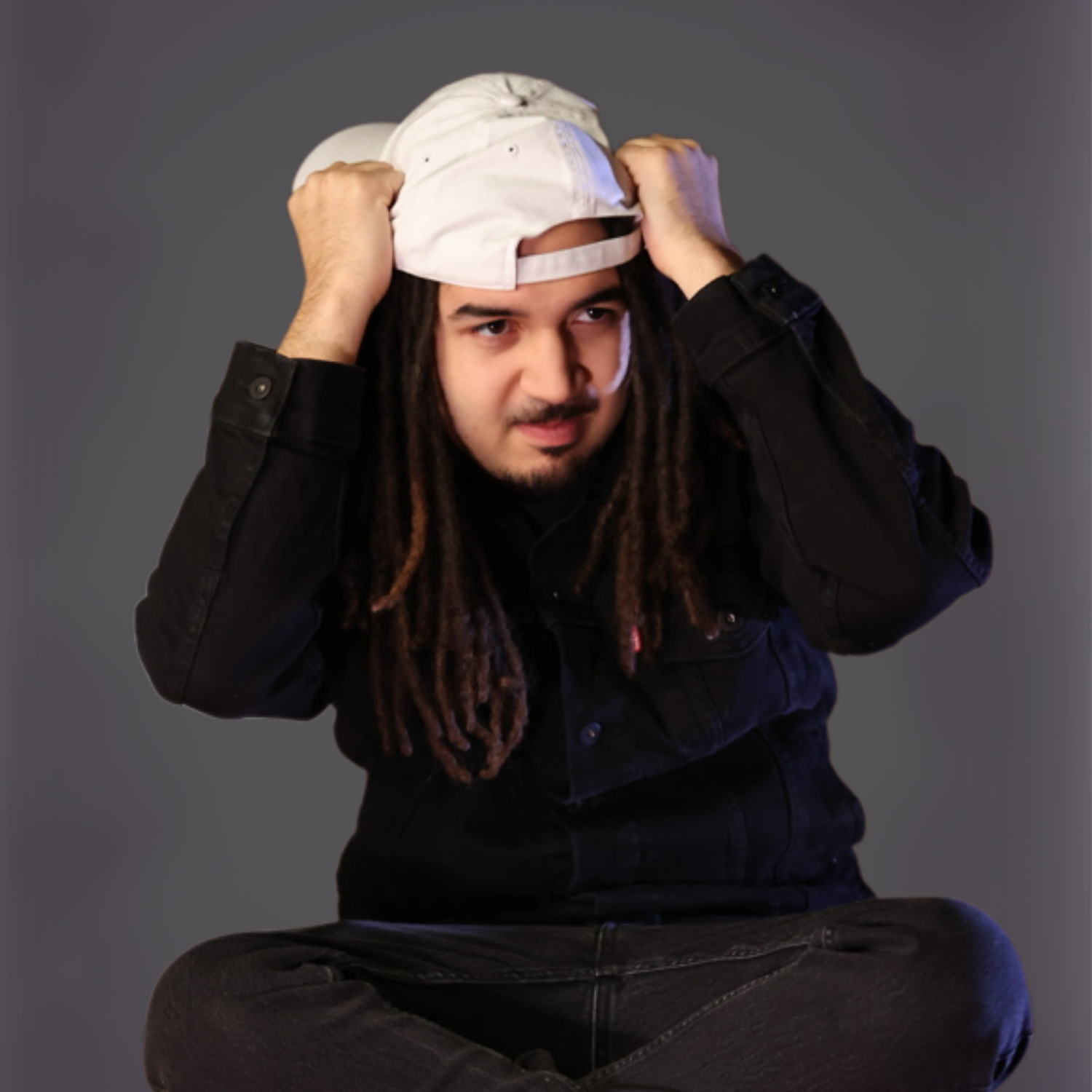 Man with locs adjusting a white baseball cap that sits too high because the hat does not have enough room for his hair volume.
