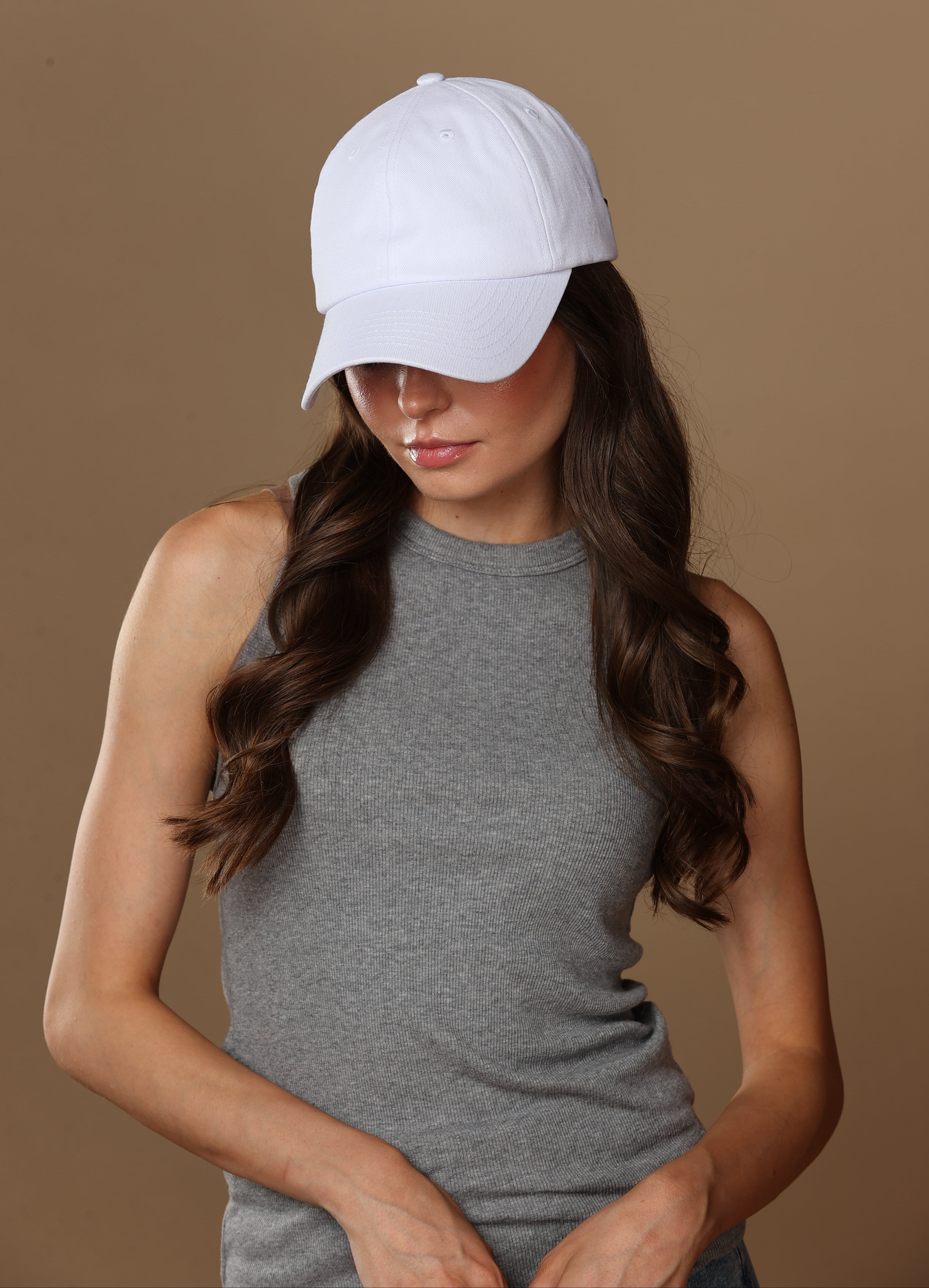 Woman with long wavy brown hair wearing a white baseball cap and gray sleeveless top against a tan studio background.