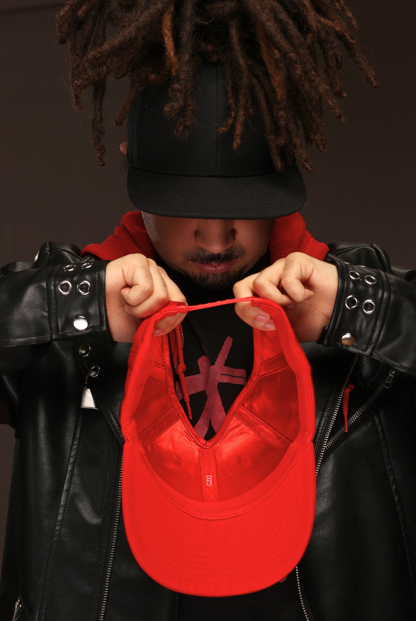 Person with locs wearing a black flat bill cap and holding a red cap toward the camera.