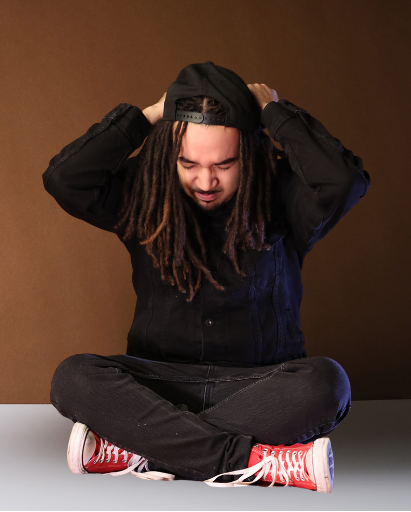 Person with long locs sitting cross-legged in a studio while adjusting a black snapback cap.