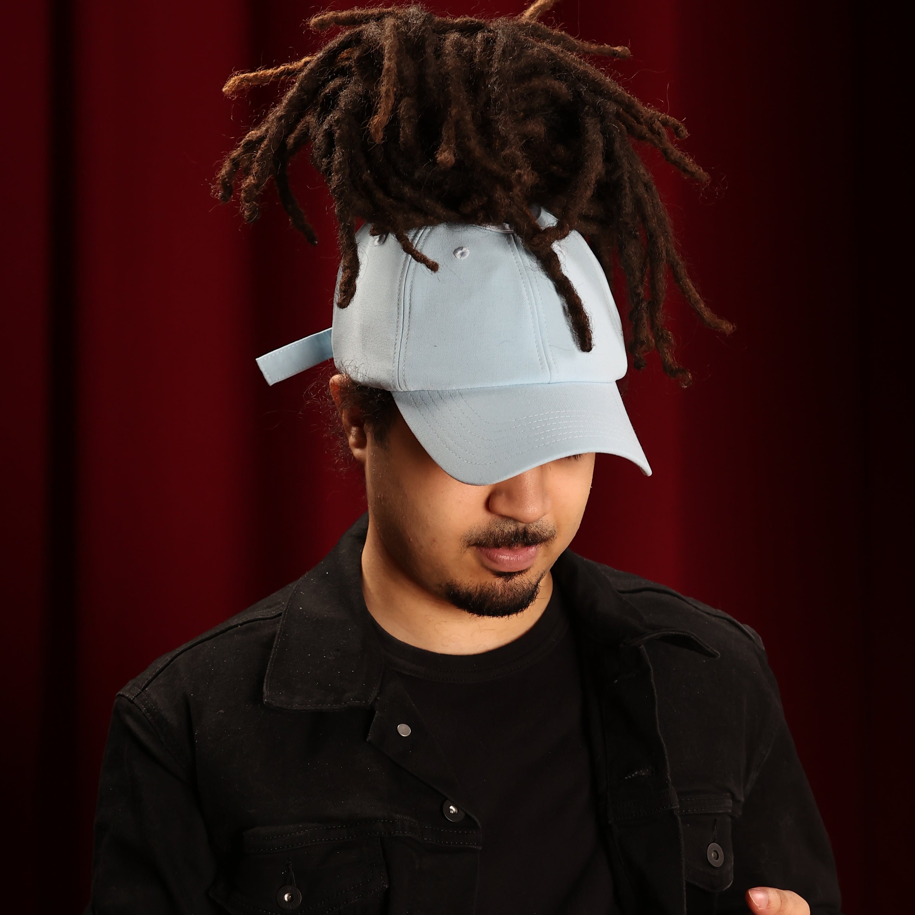 Portrait of a model wearing the baby blue Rosebloom JAX with locs pulled through the crown opening, styled with a black jacket against a red curtain backdrop.