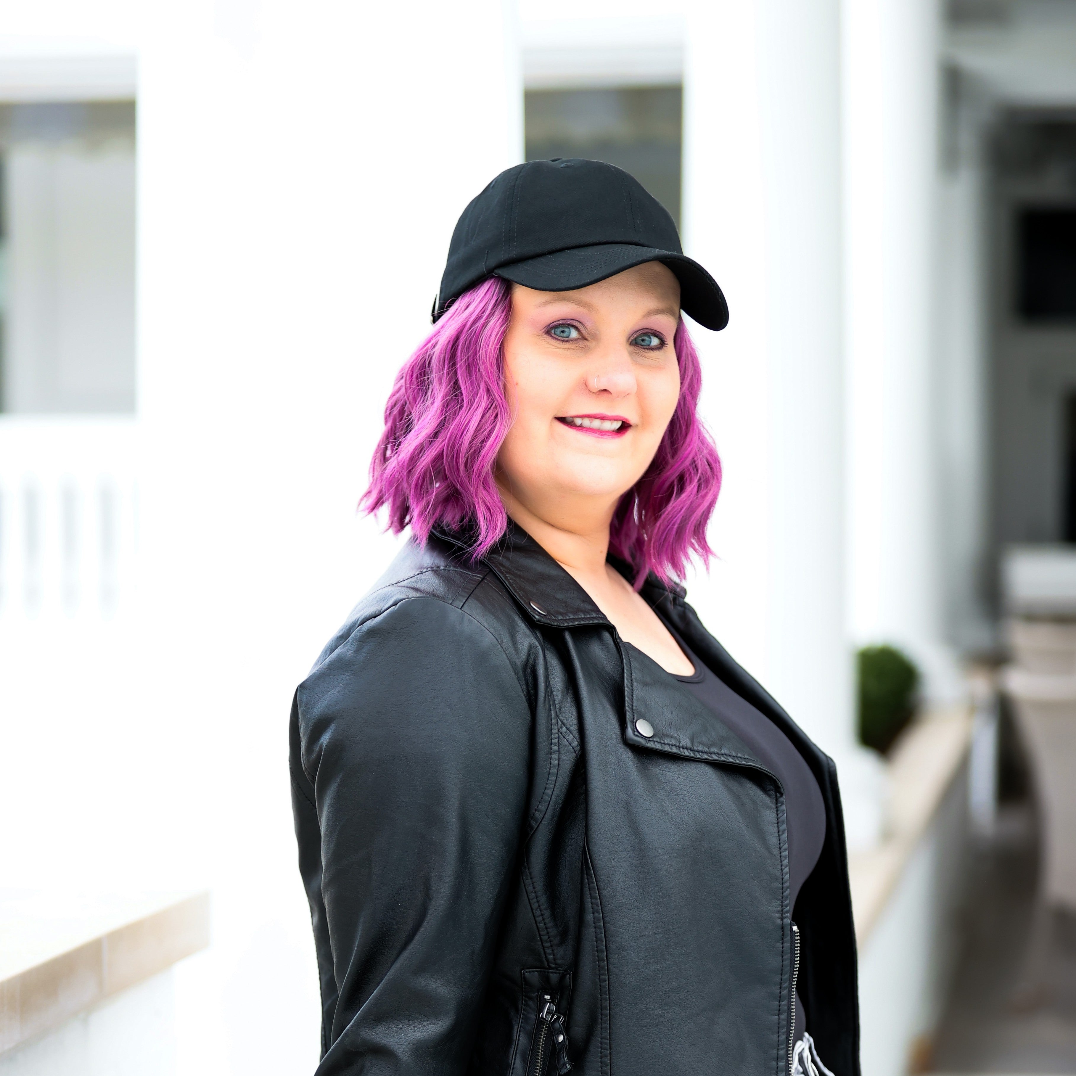 Lifestyle portrait of a model wearing a black Rosebloom Compassion Cap, styled with purple hair and a black jacket in a bright indoor setting.