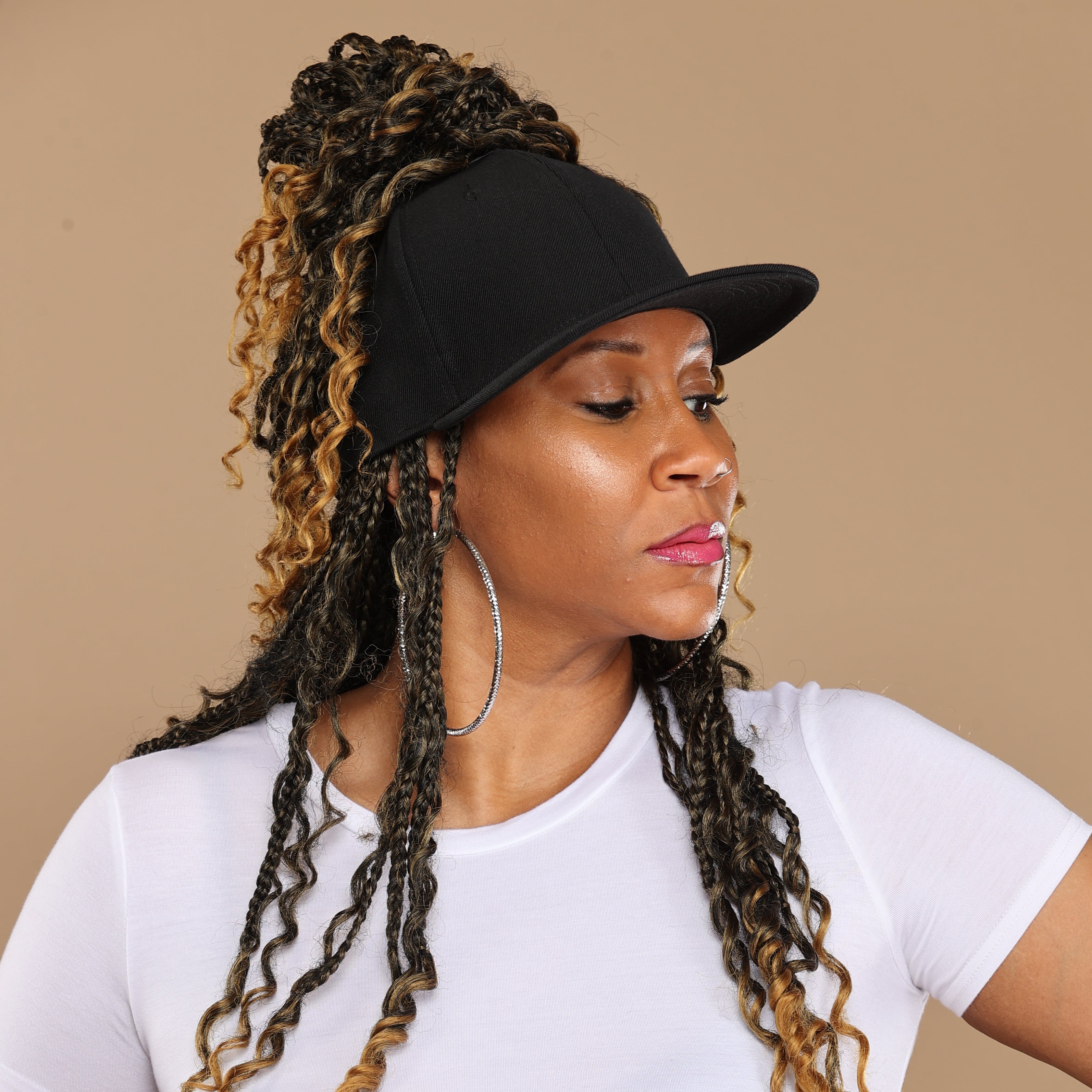 Side portrait of a model wearing a black Rosebloom GZO STRETCH with textured hair pulled through the crown opening, styled with a white top and hoop earrings against a beige studio background.