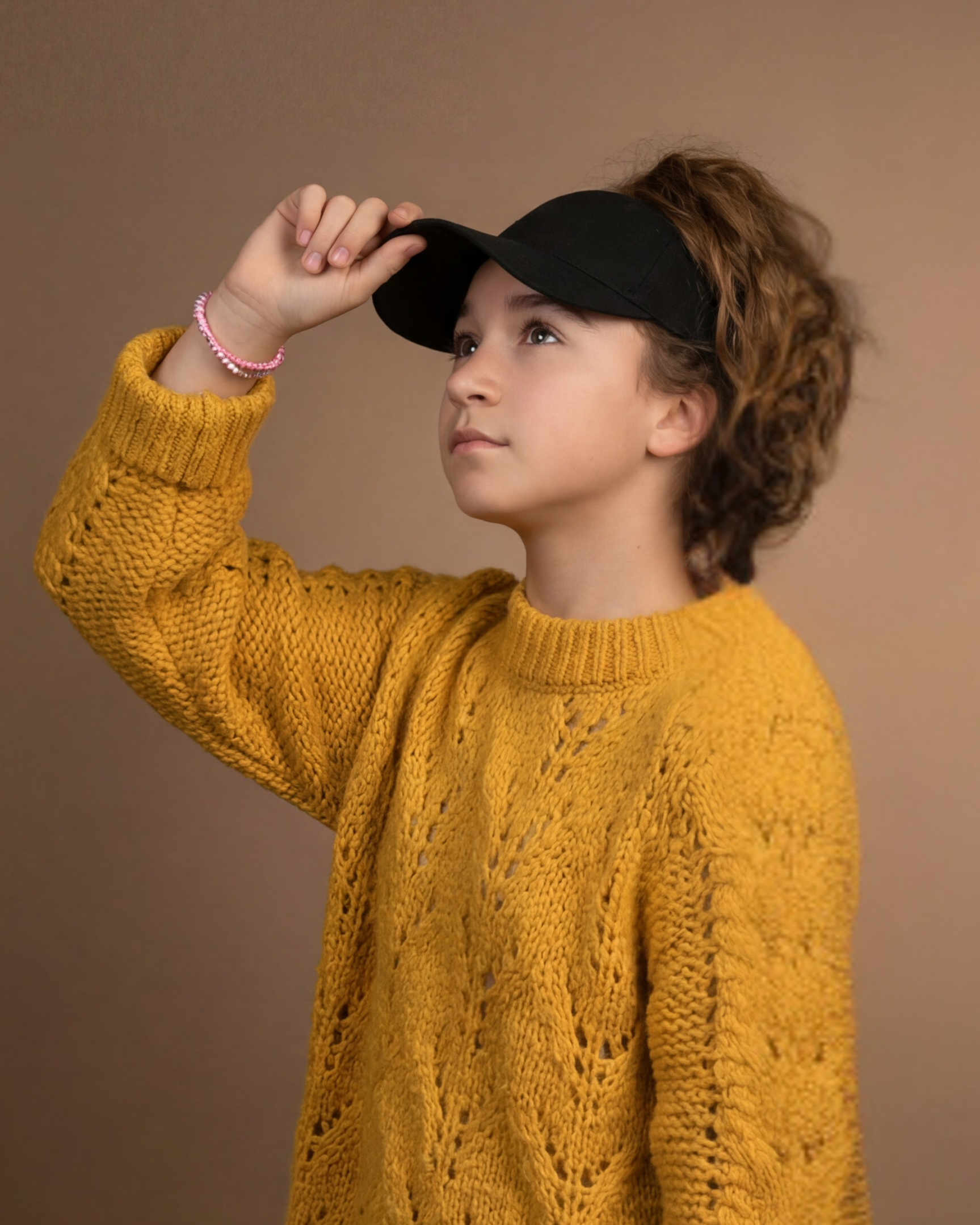 Profile portrait of a young model wearing a black Rosebloom LAZ crown-opening hat with curly hair pulled through the opening, styled with a mustard sweater against a warm studio background.