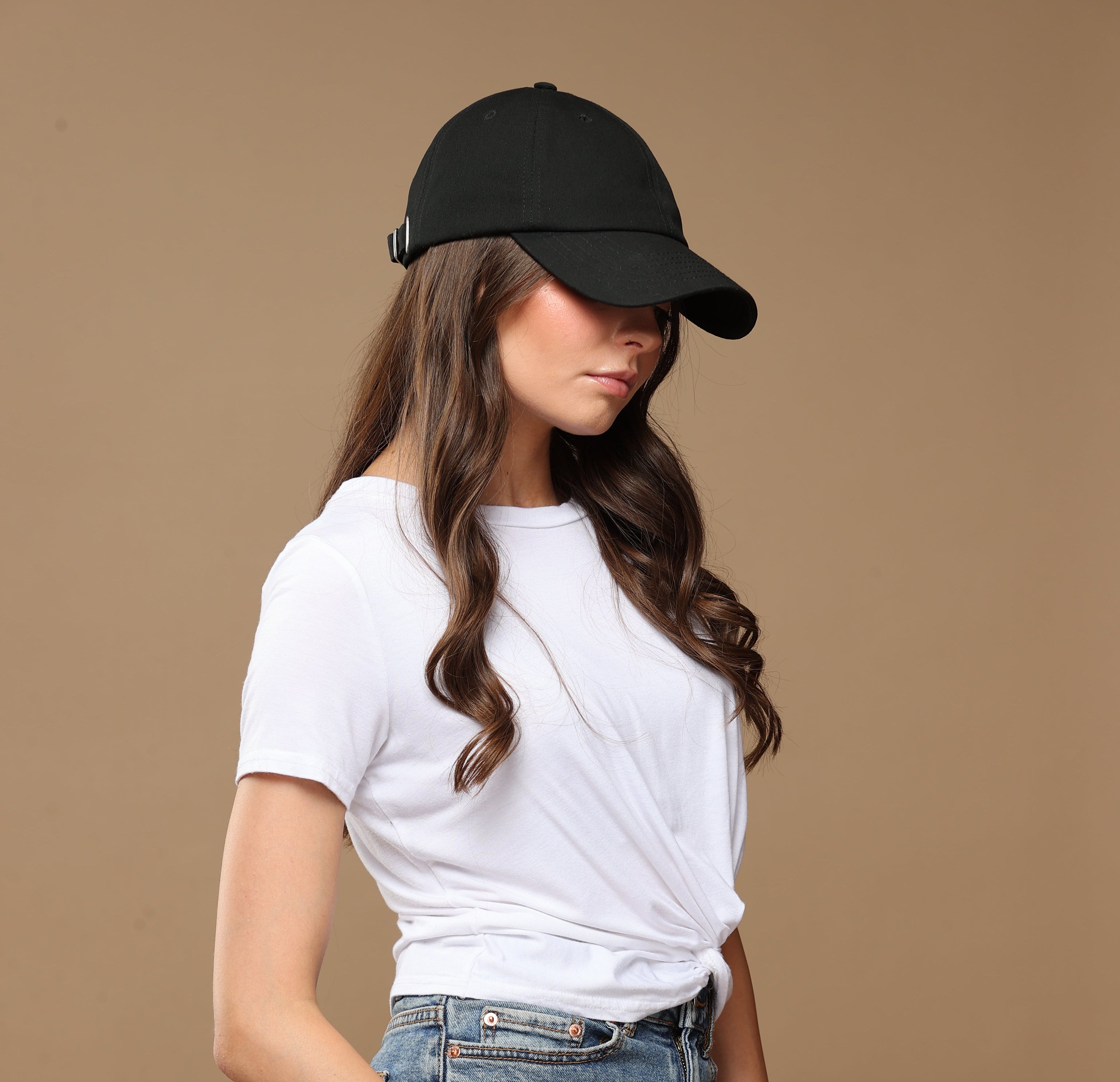 Side lifestyle portrait of a model wearing a black Rosebloom NZR women’s hat, styled with a white top and denim against a beige studio background.