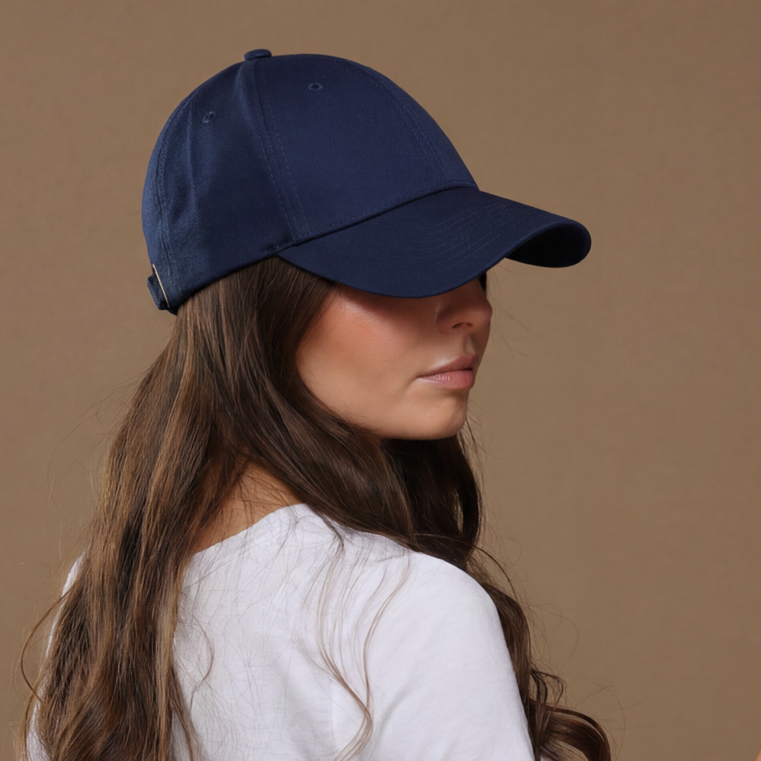 Back-side lifestyle portrait of a model wearing a blue Rosebloom NZR women’s hat, styled with a white top and denim while holding sunglasses against a beige studio background.