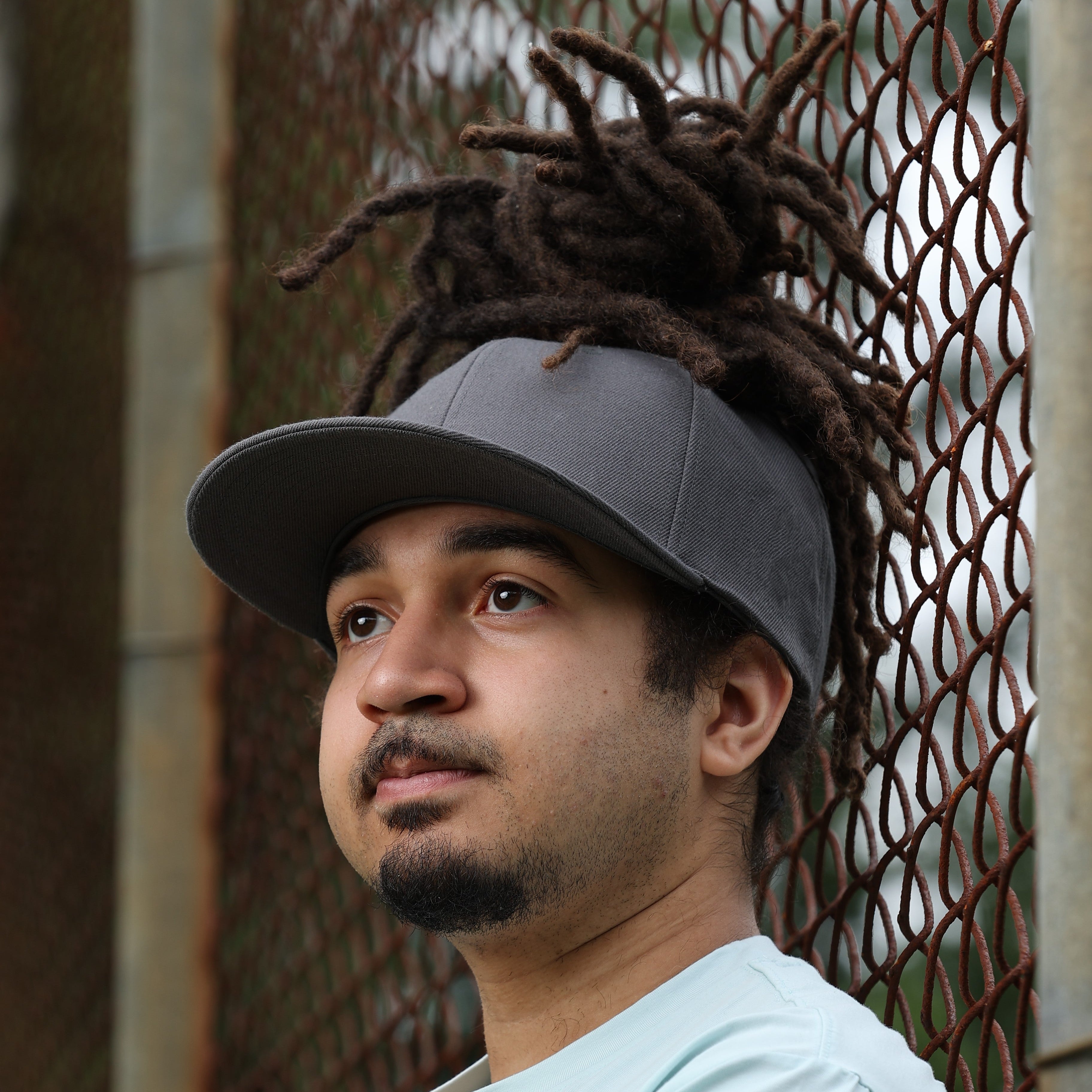 Lifestyle portrait of a model wearing a gray Rosebloom GZO STRETCH with locs pulled through the crown opening, posed outdoors against a metal fence.