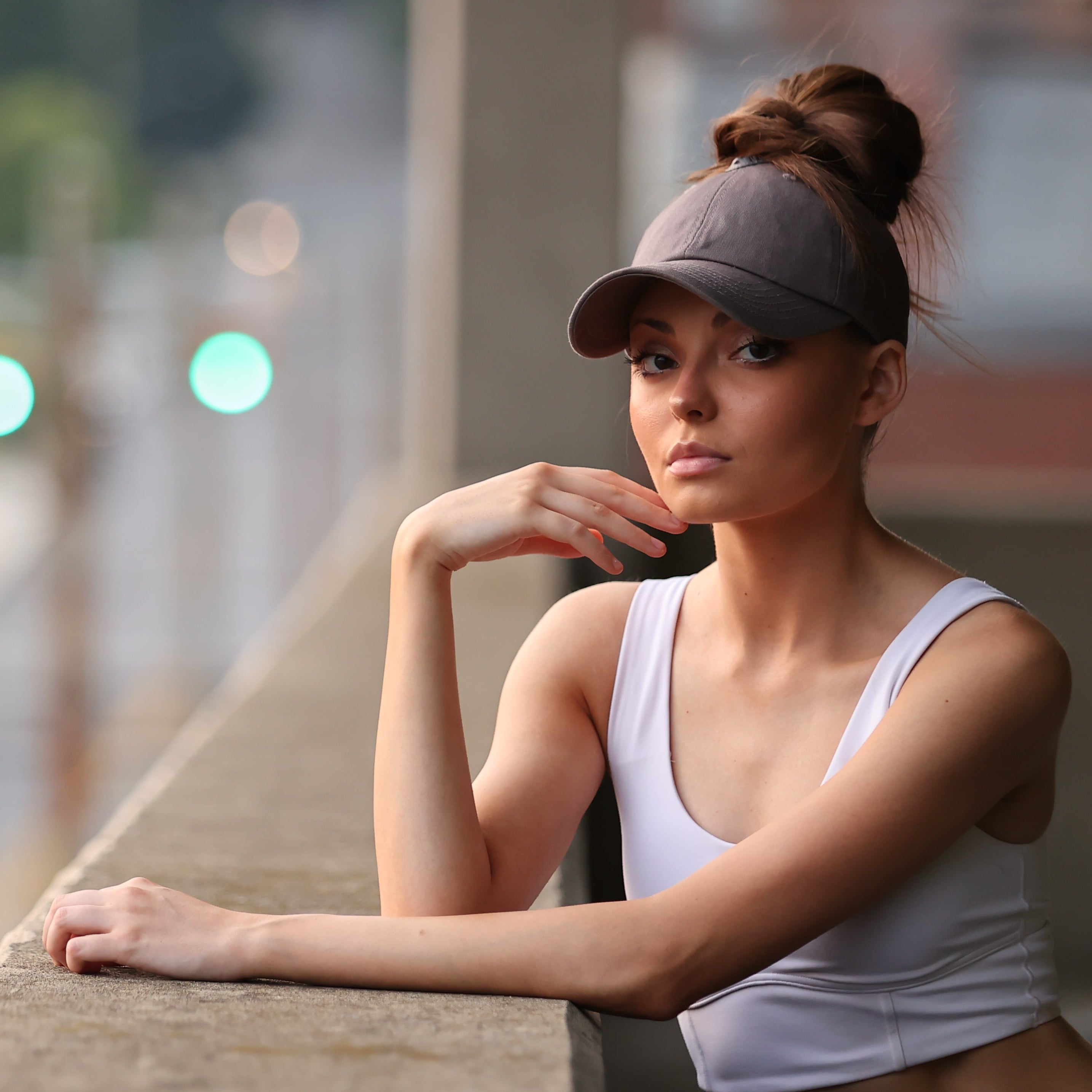 Lifestyle portrait of a model wearing a gray Rosebloom PNT high ponytail hat with hair pulled through the crown opening, leaning on a city balcony railing.