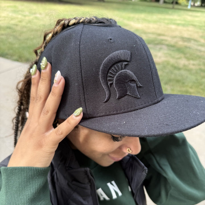 Lifestyle close-up of a model wearing the black Rosebloom Michigan State GZO SNAP with a tonal embroidered Spartan logo, styled outdoors.