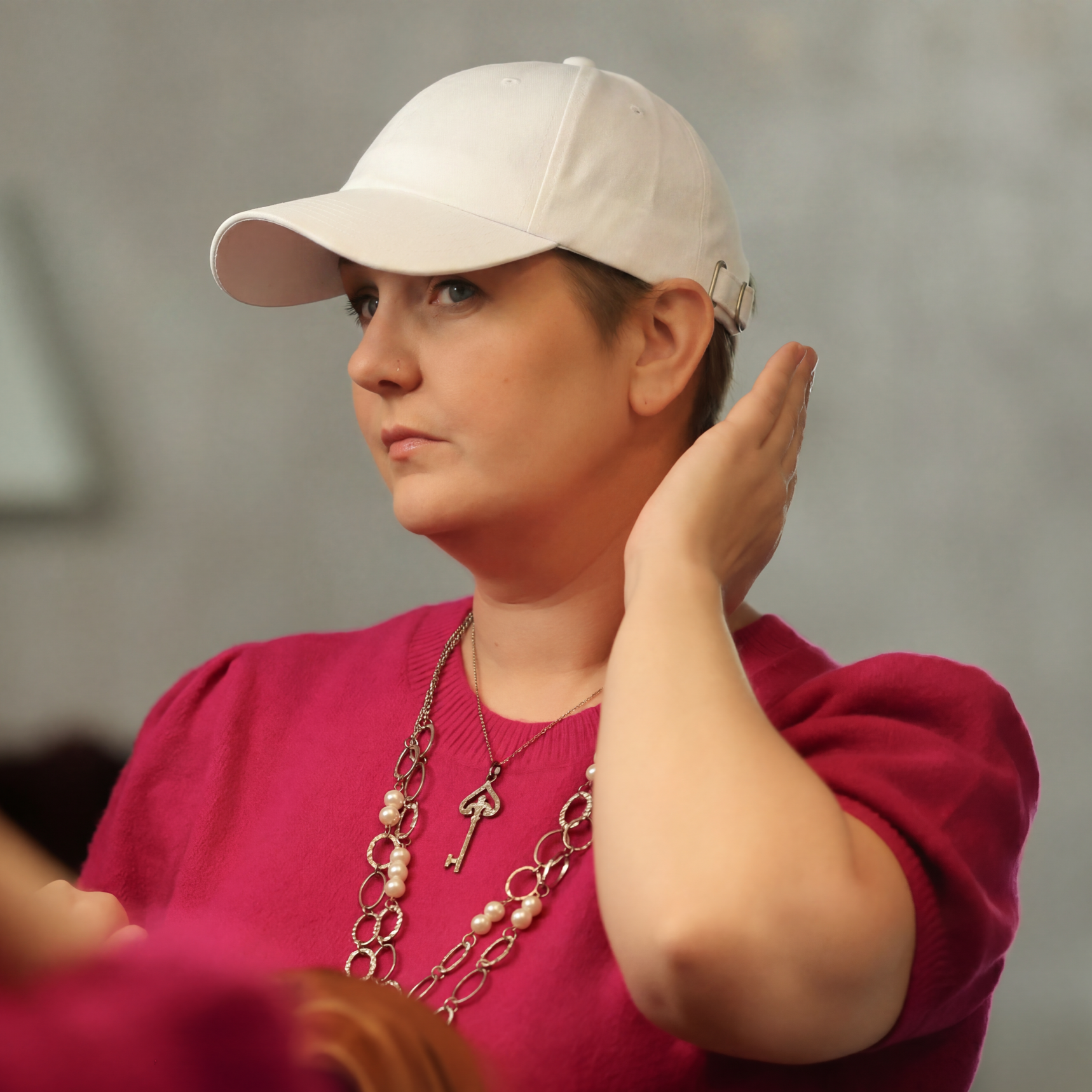 Side view of a model wearing a white Rosebloom Compassion Cap, styled with a bright pink sweater and layered necklace in a soft indoor lifestyle portrait.
