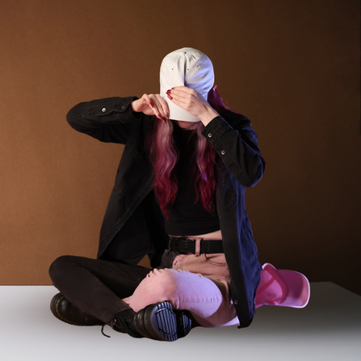 Person with pink wavy hair sitting cross-legged in a studio while adjusting a white baseball cap.